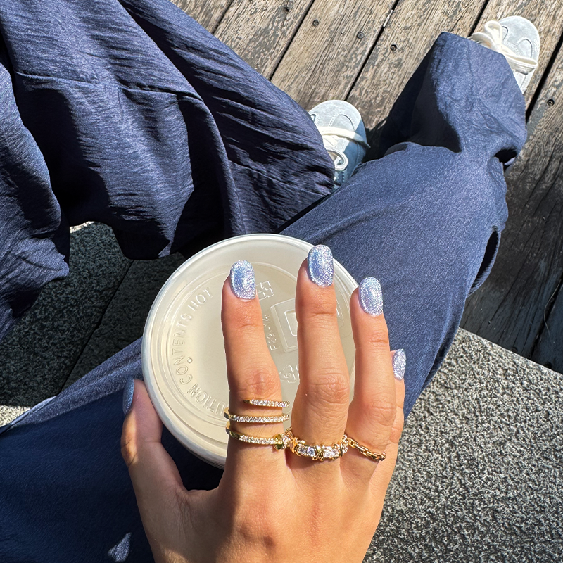 Fincloset blue cat eye handmade press on nails with shimmering galaxy effect, styled with layered gold rings while holding a coffee cup outdoors on a sunny day, chic street fashion manicure.