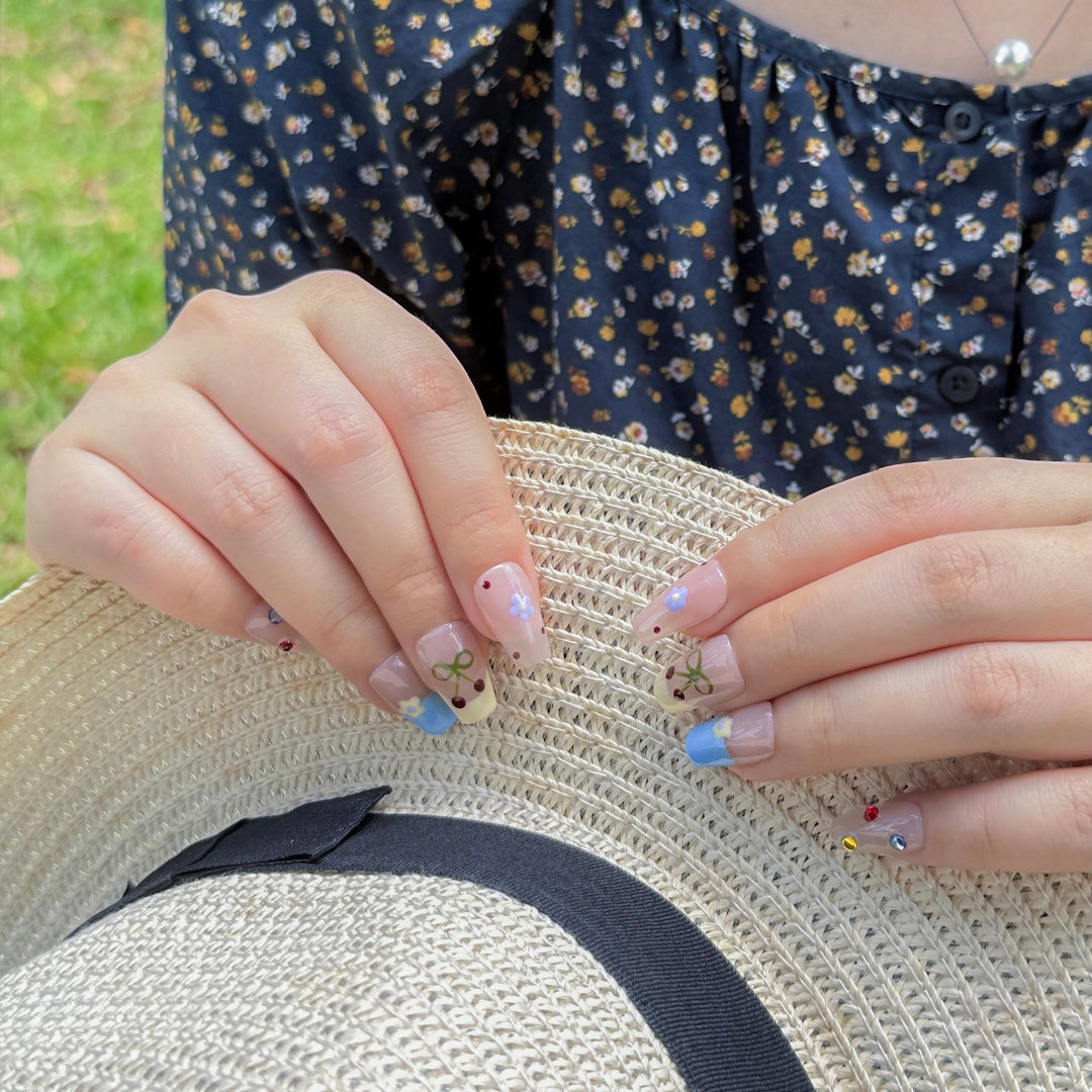 Close-up of Fincloset Dream Puff press-on nails featuring pastel nude base, blue French tips, and hand-painted floral designs, worn in a sunny outdoor picnic setting.
